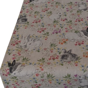 May include: A beige linen tablecloth featuring a repeating pattern of white rabbits and colourful flowers. The rabbits are in various poses, some are sitting, some are standing, and some are lying down. The flowers are in a variety of colours, including pink, purple, blue, and yellow.