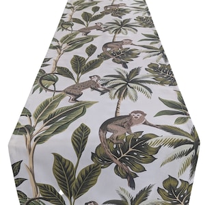 以下が含まれることがあります： サル、ヤシの木、大きな緑の葉が描かれた、熱帯のジャングル柄の白いテーブルランナーです。