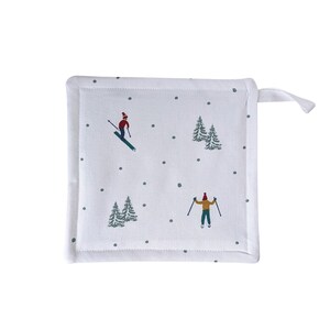 May include: White square pot holder with a winter scene print. The design features skiers, evergreen trees, and gray polka dots. The pot holder has a white border and a loop for hanging.