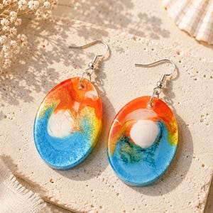 May include: A pair of oval-shaped drop earrings featuring a swirling resin design in vibrant shades of orange, bright blue, and white. Each earring is attached to a silver-toned metal hook, showcasing a smooth, glossy finish with blended colors.
