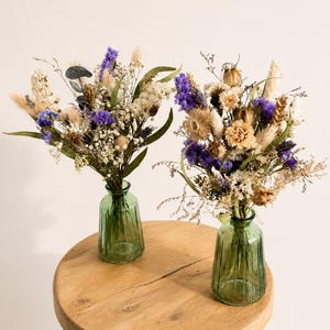May include: Two bouquets of dried flowers featuring purple, white, and beige stems, including lavender, baby's breath, and grasses. Each arrangement is displayed in a small, ribbed, translucent green glass vase with a narrow neck and base.