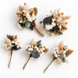 Peut inclure: Cinq boutonnières florales composées de fleurs séchées gypsophile blanches, d'herbe de la pampa beige, de feuilles d'eucalyptus vertes et de petites gousses brunes. Les tiges sont entourées de fil métallique fin ou de ficelle naturelle.