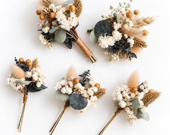 Peut inclure: Cinq boutonnières florales composées de fleurs séchées gypsophile blanches, d'herbe de la pampa beige, de feuilles d'eucalyptus vertes et de petites gousses brunes. Les tiges sont entourées de fil métallique fin ou de ficelle naturelle.