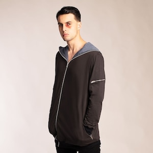 May include: A man wearing a dark grey hooded jacket with a zip closure and black trousers. The jacket has a blue hood lining and a zipped pocket on the left sleeve.