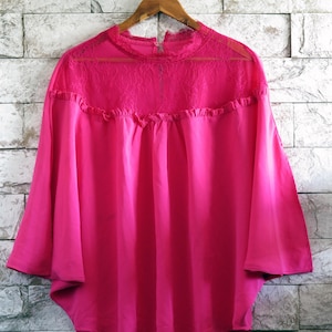 May include: A bright pink blouse with a high neckline and three-quarter sleeves. The top features a lace panel across the chest and ruffled detailing. The blouse hangs on a wooden hanger against a light gray brick wall.