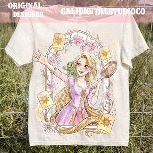 Puede incluir: Una camiseta color crema con una ilustración gráfica de un personaje rubio con vestido morado y un camaleón verde en el hombro, sosteniendo una sartén, rodeado de linternas amarillas flotantes y flores de color rosa.