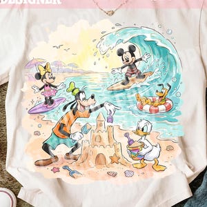 Puede incluir: Una camiseta color crema con un gráfico colorido de personajes de Disney en la playa. Mickey Mouse surfea una ola, Minnie Mouse surfea cerca, Goofy construye un castillo de arena, Pluto flota en un anillo rojo y blanco y el Pato Donald sostiene un cubo.