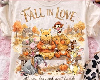 Può includere: Una t-shirt color crema con una grafica di Winnie the Pooh, Tigro, Pimpi, Ih-Oh e Tappo seduti su una panchina in un ambiente autunnale. Il testo recita "FALL IN LOVE" in alto e "with cozy days and sweet friends" nella parte inferiore.