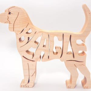 May include: A light-coloured wooden cutout of a standing dog in profile. The body of the dog is formed by the letters spelling the word BEAGLE, with the head and tail extending from the typography. The natural wood grain is visible throughout the piece.