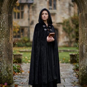 May include: A floor-length black velvet hooded cloak featuring a front closure with two dark brown toggle buttons. The garment is worn over a black outfit and is held together at the neck, providing full coverage with a deep, structured hood.
