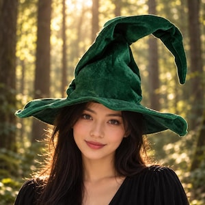 May include: A dark green velvet witch hat with a wide, flexible brim and a tall, pointed, slightly slumped crown. The fabric has a soft, textured appearance with subtle light reflections across the surface of the deep green material.