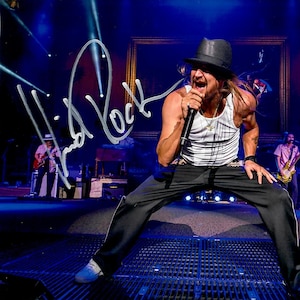 Könnte beinhalten: Ein Künstler in einem weißen Tanktop, einer schwarzen Hose und einem schwarzen Fedora-Hut, der auf einer Bühne mit blauer Beleuchtung in ein Mikrofon singt. Das Bild enthält eine weiße Signatur, die Kid Rock auf der linken Seite zeigt.
