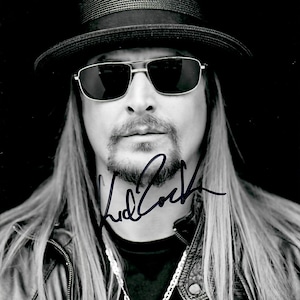 Könnte beinhalten: Ein Schwarz-Weiß-Porträt einer Person mit langem Haar, die einen dunklen Fedora-Hut, eine dunkle Sonnenbrille und eine Lederjacke trägt. Eine silberne Kette mit Anhänger ist zu sehen. Das Bild zeigt eine dunkle Tintensignatur im Brustbereich.