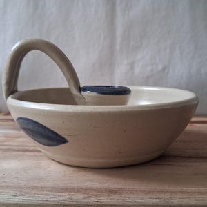 May include: A beige ceramic candle holder with a rounded bowl shape and a single arched handle. The piece features a dark blue painted leaf-like motif on the exterior side and a dark blue rim around the central candle cup opening.