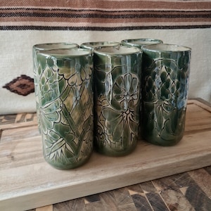 May include: A set of six cylindrical ceramic tumblers featuring a deep green glaze with carved floral and geometric patterns. Each vessel has a smooth, glossy finish and a consistent tall, straight-sided shape with a rounded base rim.
