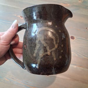 May include: A dark brown ceramic pitcher with a glossy, speckled glaze and a single handle. The body features a lighter, abstract, hand-painted circular design on the front, with a small pouring spout at the top rim of the vessel.