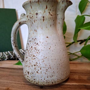 May include: A ceramic pitcher with a speckled cream and brown glaze. The vessel features a wide, flared rim, a curved handle on the side, and a distinct pouring spout. The surface is covered in irregular dark brown spots over a light base.