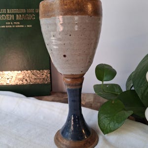 May include: A ceramic goblet featuring a speckled off-white bowl with a brown rim. The stem and base are finished in a deep blue glaze with brown accents at the bottom. The surface of the bowl shows subtle horizontal ridges from the pottery process.