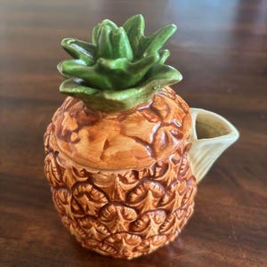 May include: A ceramic pineapple-shaped creamer or pitcher with a removable lid. The body features a textured brown and orange pineapple pattern, while the lid is shaped like green leaves. A small cream-coloured spout extends from the side.