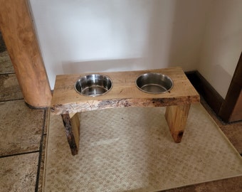 May include: A rustic wooden dog food stand with two stainless steel bowls. The stand has a natural wood finish and is elevated on two legs. The stand is placed on a beige mat with a paw print pattern.