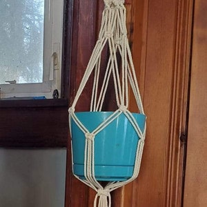May include: A white macrame plant hanger with two pots. The top pot is turquoise blue and the bottom pot is a light purple. The hanger is hanging from a hook on a wooden wall.