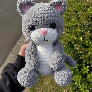 May include: A handmade crochet cat plush toy made from soft grey and white yarn. The cat features large black plastic eyes, a small pink stitched nose, a white muzzle, and pink inner ears. It has a rounded head, a white belly, and a long tail.