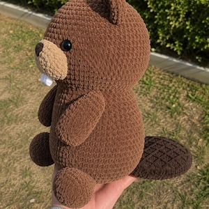 Puede incluir: Un castor de peluche hecho de hilo marrón con un acabado de ganchillo texturizado. Presenta un hocico de color tostado claro, un único diente blanco visible, ojos negros pequeños, una nariz marrón oscura y una cola plana con patrón de rejilla.