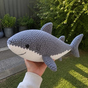 May include: A handmade crochet shark plush toy featuring a textured grey upper body and a smooth white underbelly. The shark has a prominent dorsal fin, side fins, a tail fin, and a small black button eye with a stitched smile on its face.
