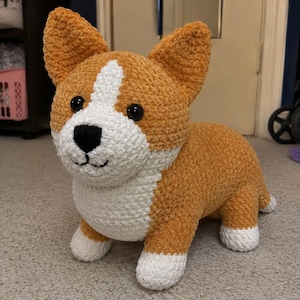 Könnte beinhalten: Ein handgefertigtes Häkel-Stofftier in Form eines Corgi-Hundes. Das Plüschtier hat eine goldbraune und weiße Garnstruktur mit markanten spitzen Ohren, schwarzen Kunststoffaugen, einer schwarzen gestickten Nase sowie weißer Brust und Pfoten.