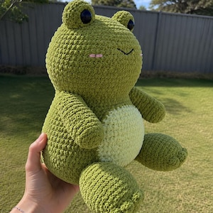 Könnte beinhalten: Ein handgefertigtes Häkel-Frosch-Stofftier mit einem strukturierten olivgrünen Körper, einem helleren, blassgrünen runden Bauch, zwei markanten dunklen runden Augen und kleinen gestickten rosa horizontalen Linien auf den Wangen als Erröten.