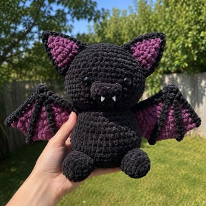 May include: A plush crochet bat toy made from soft black yarn with purple accents on the inner ears and wings. The bat features two round black eyes and two small white fangs protruding from its mouth, with a rounded body and outstretched wings.