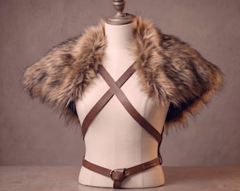 May include: A brown faux fur shoulder wrap paired with a brown leather harness featuring crisscross straps across the chest and a waist belt with a metal ring buckle. The accessory is displayed on a neutral-toned mannequin torso.