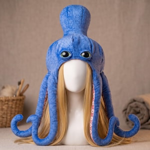May include: A blue octopus-shaped hat featuring a textured scale pattern on the exterior, two large dark eyes, and multiple dangling tentacles with pink undersides. The hat is displayed on a mannequin head with long blonde hair.