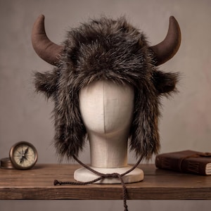 Op de afbeelding: Een bruine imitatiebontmuts met twee grote, gebogen, bruine hoorns aan de zijkanten. De muts heeft lang, ruig donkerbruin bont over de kruin en de oorflappen, met dunne gevlochten bruine koordjes die aan de onderkant naar beneden hangen.