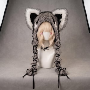 May include: A faux fur hood featuring large, pointed animal ears with white and grey colouring. The hood has long, dangling side panels laced with thin black ribbons that tie at the ends. It is displayed on a mannequin with blonde hair.