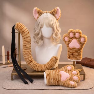 May include: A set of orange and brown striped faux fur cat accessories including a headband with ears, a long tail with black straps and metal rings, and two paw-shaped gloves with pink pads, displayed on a white mannequin head and stands.