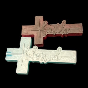 May include: Two cross-shaped decorative items. The top cross is a deep reddish-brown with the word faith written in a cursive script. The bottom cross is a pale, iridescent light blue with the word blessed written in a cursive script.