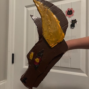 May include: A brown cardboard dragon mask featuring a large, textured yellow horn on top and painted red and yellow eyes. The mask is constructed from layered corrugated cardboard with visible seams and a hand-painted finish.