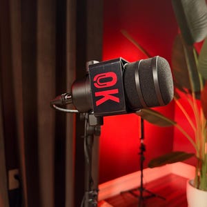 May include: A black microphone mounted on a stand, featuring a rectangular black flag attached to the body. The flag displays the letters OK in a bold, bright red font. The microphone has a black foam windscreen and a black cable extending from the base.
