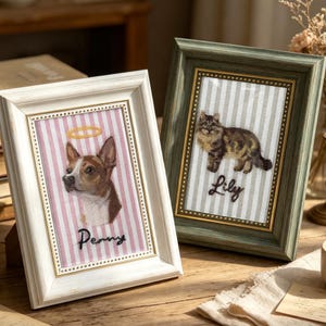 May include: Two rectangular picture frames containing embroidered portraits. The white frame features a dog with a halo and the name Penny. The green frame features a cat and the name Lily. Both portraits are set against striped fabric backgrounds.