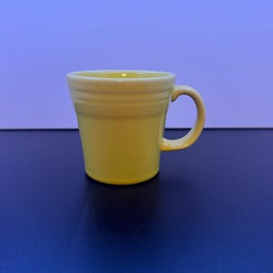 May include: A bright yellow ceramic mug featuring a flared rim and a series of three horizontal embossed rings around the upper portion of the body. The mug has a curved handle on the right side and a smooth, glossy finish throughout the exterior.