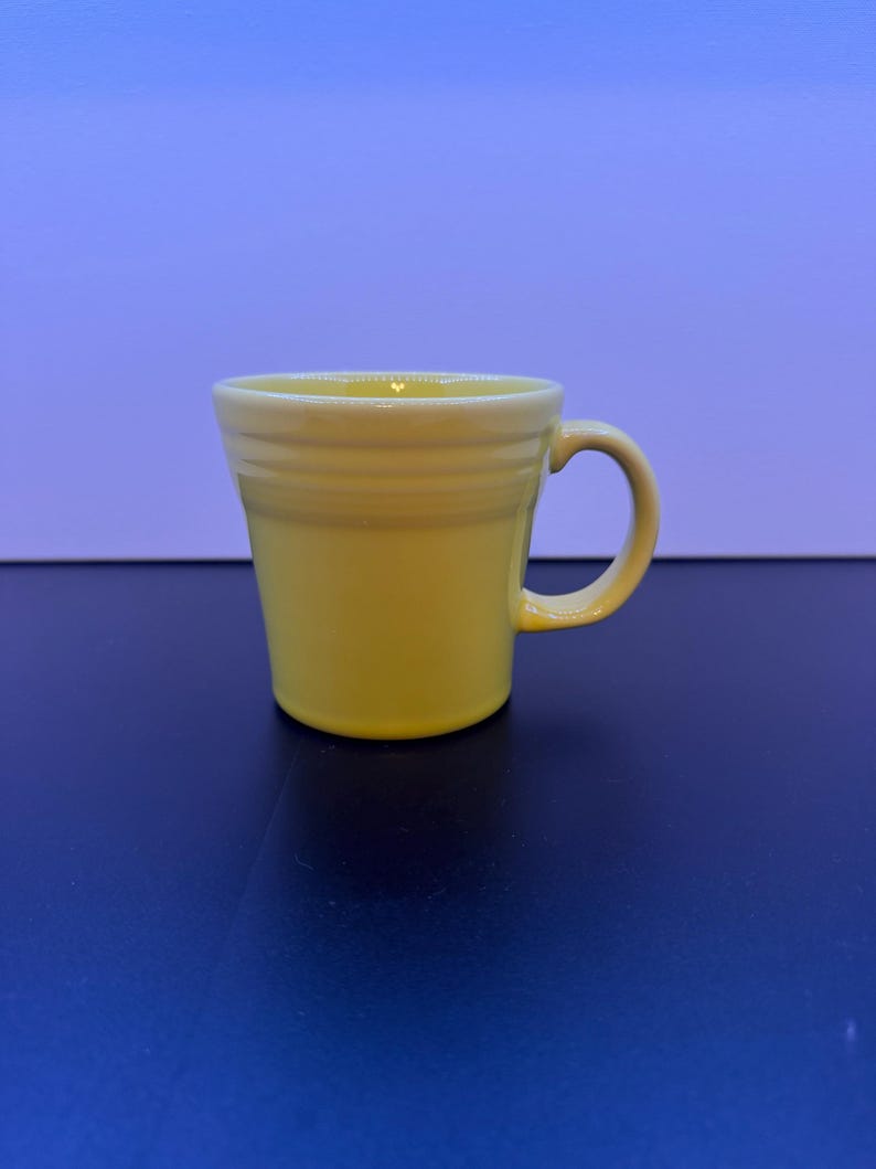 May include: A bright yellow ceramic mug featuring a flared rim and a series of three horizontal embossed rings around the upper portion of the body. The mug has a curved handle on the right side and a smooth, glossy finish throughout the exterior.