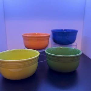May include: A set of four small ceramic bowls in vibrant colors including lime green, bright orange, deep blue, and a muted green. Each bowl features a smooth, rounded shape with a series of three horizontal rings embossed around the upper rim.