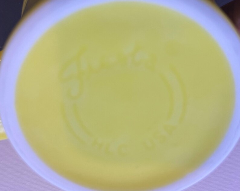 May include: The underside of a bright yellow ceramic mug featuring the embossed script text Fiesta and the letters HLC USA. The circular base has a smooth, glossy finish with a slightly raised rim around the perimeter of the bottom surface.
