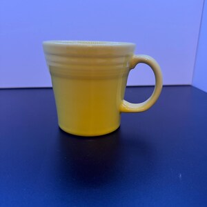 May include: A bright yellow ceramic mug featuring a smooth, glossy finish and a tapered cylindrical body. The mug includes a curved handle on the right side and three distinct horizontal ridges encircling the upper portion of the exterior wall.