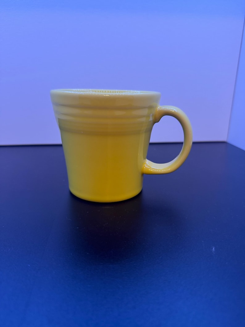 May include: A bright yellow ceramic mug featuring a smooth, glossy finish and a tapered cylindrical body. The mug includes a curved handle on the right side and three distinct horizontal ridges encircling the upper portion of the exterior wall.