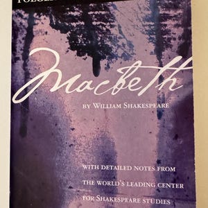 May include: A paperback book titled Macbeth by William Shakespeare, Folger Shakespeare Library Updated Edition. The cover features a purple and black abstract watercolor design with white text. It includes detailed notes from the world's leading center for Shakespeare studies, edited by Barbara A. Mowat and Paul Werstine.