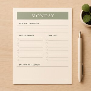 May include: A cream-colored daily planner sheet with a sage green header labeled MONDAY. The layout includes sections for Morning Intention, Top Priorities, Task List, and Evening Reflection with checkboxes and lines. A white pen sits beside the paper.