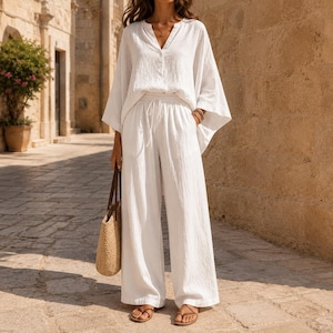 May include: A white two-piece set consisting of a long-sleeved V-neck top and wide-leg trousers with an elastic drawstring waistband. The outfit is made of a textured fabric and is paired with a woven straw tote bag and brown leather sandals.