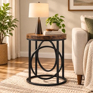May include: A round side table with a dark wood grain top and a black metal frame featuring circular accents. The table supports a conical lamp with a white shade, a small potted green plant, and two stacked books titled Home and Modern Living.
